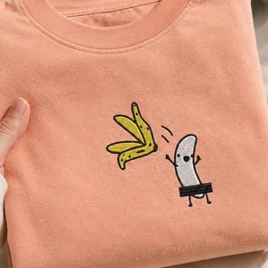 May include: A peach-colored crewneck sweatshirt featuring an embroidered graphic of a white banana character peeling off its yellow skin. The character has a simple face and a black rectangular bar covering its midsection, with thin black limbs.