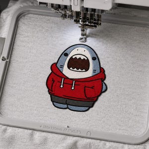 Pu&ograve; includere: Un telaio per macchina da ricamo che tiene un tessuto grigio chiaro con il disegno ricamato di uno squalo cartoon che indossa una felpa rossa con cappuccio e pantaloncini scuri. Il telaio riporta le dimensioni di 180x130mm e 7.1x5.1 pollici.