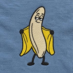 Peut inclure: Un tissu bleu clair avec une broderie représentant un personnage de banane. La banane est de couleur crème avec un visage souriant, tenant ouverte sa peau jaune vif comme un manteau, révélant de fins bras et jambes noirs.