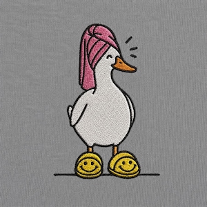 Könnte beinhalten: Ein gesticktes Motiv auf grauem Stoffhintergrund, das eine weiße Ente mit einem um den Kopf gewickelten rosa Handtuch zeigt. Die Ente hat einen orangefarbenen Schnabel und trägt gelbe Hausschuhe mit schwarzen Smiley-Gesichtern auf der Vorderseite.