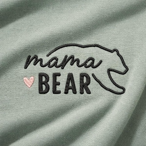 May include: Close-up of a light green fabric with the embroidered text "mama BEAR" in black, with a pink heart. A black bear outline is above the word "BEAR". The fabric has a textured appearance.