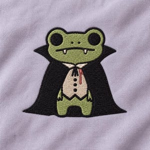 Puede incluir: Una tela de color morado claro con un parche bordado de una rana verde vestida de vampiro. La rana tiene ojos negros, colmillos blancos y lleva una capa negra sobre un chaleco color crema con tres botones negros y una gota de sangre roja.