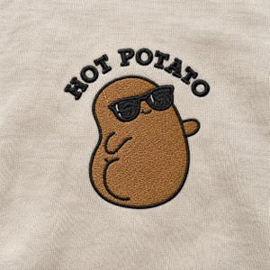 May include: A beige sweatshirt featuring an embroidered graphic of a brown potato character wearing black sunglasses. The potato has a small smile and is waving one arm. Above the character, the text HOT POTATO is embroidered in black.