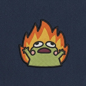 Op de afbeelding: Geborduurde patch met een groen stripfiguur met een verbaasde uitdrukking, omgeven door oranje en gele vlammen. De patch heeft een zwarte rand en staat op een marineblauwe achtergrond.