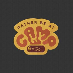 Peut inclure: Un écusson jaune et orange avec les mots "RATHER BE AT CAMP" dans une police stylisée. Le mot "CAMP" est dans une police plus grande et arrondie. Une petite illustration d'un feu de camp et d'une bûche se trouve au bas de l'écusson.