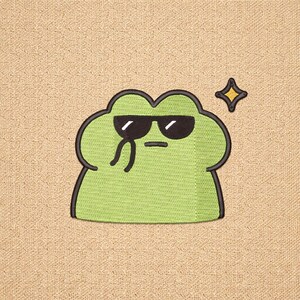 May include: A green embroidered patch featuring a cartoon frog wearing black sunglasses and a black outline. A yellow star is next to the frog. The patch is on a tan fabric background.
