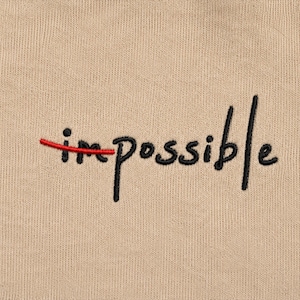 May include: A beige fabric surface featuring black embroidered text that reads "impossible". A horizontal red embroidered line strikes through the first two letters, "im", visually transforming the word to read as "possible".
