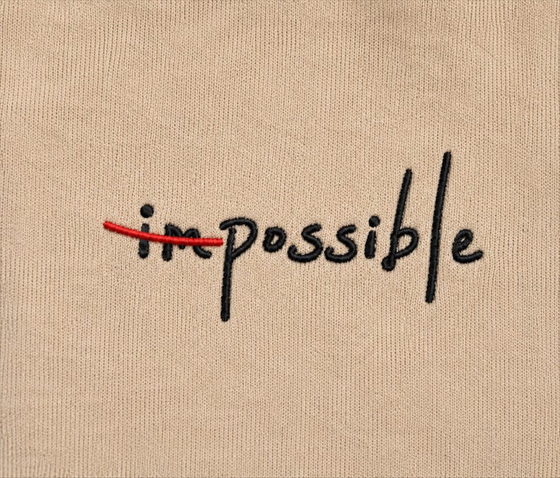 May include: A beige fabric surface featuring black embroidered text that reads "impossible". A horizontal red embroidered line strikes through the first two letters, "im", visually transforming the word to read as "possible".