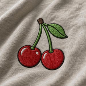 Könnte beinhalten: Ein beigefarbenes Kleidungsstück aus Stoff mit einer Kirschstickerei. Das Motiv zeigt zwei leuchtend rote Kirschen mit weißen Glanzlichtern, die über grüne Stiele mit einem braunen Stielansatz verbunden sind, ergänzt durch ein grünes Blatt.