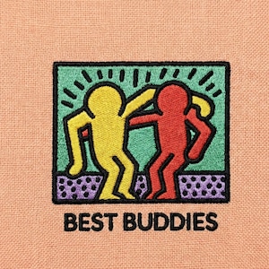 Könnte beinhalten: Gestickter Aufnäher mit zwei stilisierten Figuren in Gelb und Rot, die sich in einem grünen Rechteck umarmen. Der Aufnäher ist schwarz umrandet, darunter steht "BEST BUDDIES". Der Hintergrund ist ein pfirsichfarbener Stoff.
