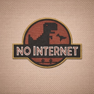 Puede incluir: Parche bordado con un dinosaurio negro pixelado y una criatura voladora dentro de un círculo rojo. El texto "NO INTERNET" está en blanco sobre un rectángulo negro, con un borde dorado. Dos cactus en la parte inferior. Fondo beige.
