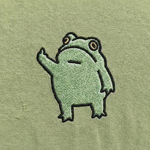 May include: A green embroidered frog with a black outline giving the middle finger. The frog has yellow eyes and is on a light green background. The frog is a cartoon-like design.