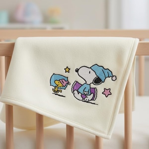 May include: Cream-colored baby blanket with an embroidered Snoopy and Woodstock design. Snoopy is wearing a blue nightcap and Woodstock is wearing a pink nightcap. The blanket is draped over a wooden crib.