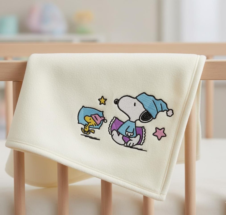 May include: Cream-colored baby blanket with an embroidered Snoopy and Woodstock design. Snoopy is wearing a blue nightcap and Woodstock is wearing a pink nightcap. The blanket is draped over a wooden crib.