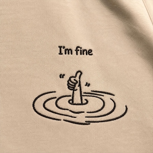 Puede incluir: Una prenda de tela beige con bordado negro. El diseño muestra una mano emergiendo del agua haciendo un gesto de pulgar hacia arriba, rodeada de ondas circulares. Sobre el gráfico, el texto "I'm fine" está bordado en color negro.