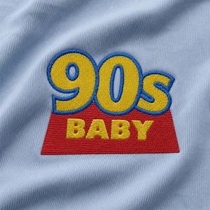 Pode incluir: Um tecido azul claro com um emblema bordado com o texto 90s BABY. O 90s é costurado em amarelo com um contorno azul, enquanto a palavra BABY é costurada em amarelo sobre um fundo trapezoidal vermelho com uma borda azul.