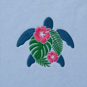May include: Embroidered design of a sea turtle with a teal body and flippers. The turtle's shell is composed of green leaves and pink hibiscus flowers. The background is a light blue fabric.
