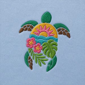 May include: Embroidered sea turtle design in green, brown, yellow, pink, and turquoise on a light blue background. The turtle features a sunset, flowers, and tropical leaves. The design is likely for a shirt or other apparel item.