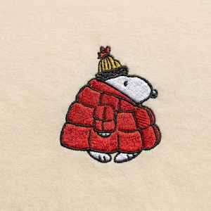 Könnte beinhalten: Gestickter Snoopy mit roter Steppjacke und gelb-schwarzer Strickmütze. Der weiß-schwarze Snoopy ist auf cremefarbenem Hintergrund gestickt. Das Design ist eine charmante Darstellung des beliebten Zeichentrickcharakters.