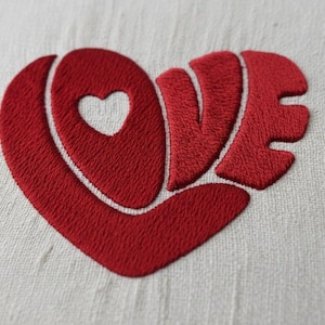Puede incluir: Un corazón rojo bordado con la palabra "LOVE" cosida en el diseño. La forma de corazón tiene un corazón más pequeño recortado en el centro. El bordado está sobre un fondo de tela blanca.