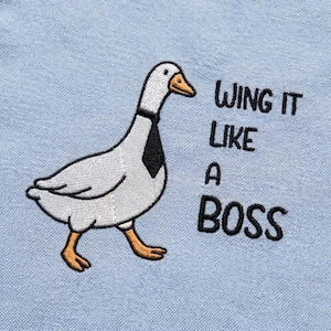 Pode incluir: Ganso bordado usando uma gravata preta, com o texto "WING IT LIKE A BOSS" em preto. O ganso é branco e cinza com pés e bico laranja, em um fundo azul claro.