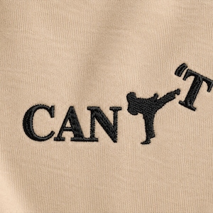 Pode incluir: Uma peça de tecido bege com texto bordado em preto que diz CAN seguido pela silhueta de uma pessoa realizando um chute de artes marciais, e terminando com a letra T, formando a palavra CAN'T com um gráfico de chute.
