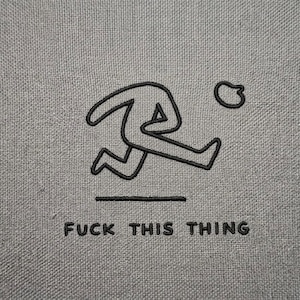 May include: Gray fabric with a black embroidered design of a running figure and a small circle. Below the figure, the text "FUCK THIS THING" is embroidered in black.