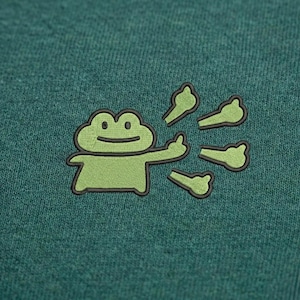 May include: A green embroidered patch featuring a cartoon frog giving a thumbs up. The frog is light green with a black outline, and the thumbs up gestures are also light green with black outlines. The background is a textured teal fabric.