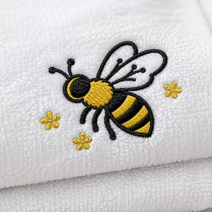 May include: White towel with an embroidered bee design. The bee is yellow and black with black outlines, and is surrounded by three small yellow flower designs. The towel is folded, showing a textured surface.