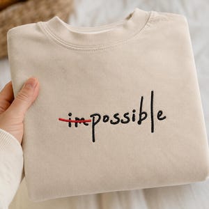 May include: A beige crewneck sweatshirt featuring black embroidered text that reads "impossible" with the "im" portion crossed out by a horizontal red embroidered line. The garment has a ribbed collar and long sleeves, presented in a folded style.