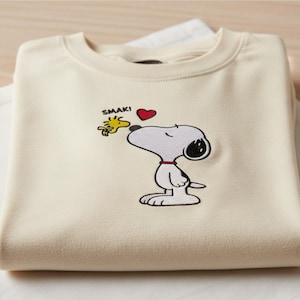 May include: Cream-colored sweatshirt featuring an embroidered Snoopy design with Woodstock and a red heart. The word "SMAK!" is above the characters. The sweatshirt is folded and resting on a white surface.
