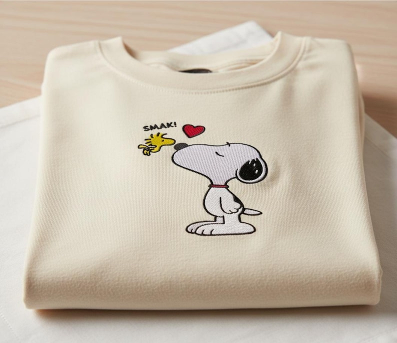May include: Cream-colored sweatshirt featuring an embroidered Snoopy design with Woodstock and a red heart. The word "SMAK!" is above the characters. The sweatshirt is folded and resting on a white surface.