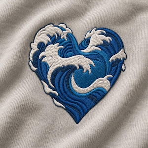 Puede incluir: Una prenda de tela gris claro con un bordado en forma de corazón. El bordado representa olas oceánicas estilizadas en tonos azul oscuro, azul medio y blanco, con las crestas de las olas formando la silueta del corazón.