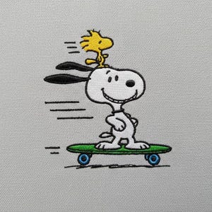 May include: Embroidered image of Snoopy skateboarding with Woodstock on his head. Snoopy is white with black ears and a black nose, riding a green skateboard with blue wheels. Woodstock is yellow. The image is on a light gray background.