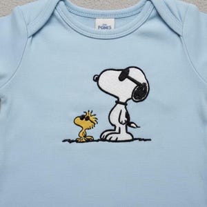May include: A light blue baby onesie featuring an embroidered image of Snoopy wearing sunglasses and Woodstock. The onesie has a round neckline and short sleeves. The brand name "POMIS" is printed on a small tag.