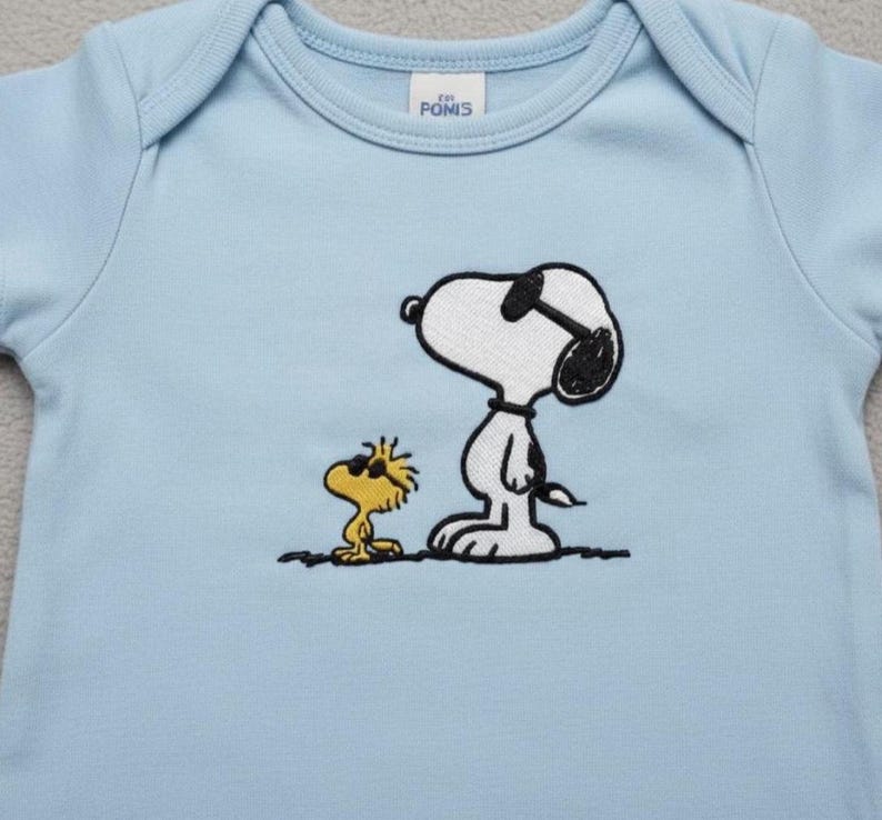 May include: A light blue baby onesie featuring an embroidered image of Snoopy wearing sunglasses and Woodstock. The onesie has a round neckline and short sleeves. The brand name "POMIS" is printed on a small tag.
