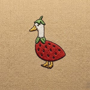 Könnte beinhalten: Gesticktes Erdbeer-Ente-Design auf beigem Stoff. Die Ente hat einen weißen Hals, einen gelben Schnabel und trägt ein rotes Erdbeerkostüm mit grünen Blättern auf dem Kopf und um den Hals. Die Füße der Ente sind gelb.