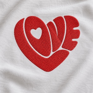 May include: A white fabric garment featuring a large, bold, red embroidered design in the shape of a heart. The embroidery incorporates the word LOVE, with the letter O represented by a smaller, negative-space heart shape within the design.