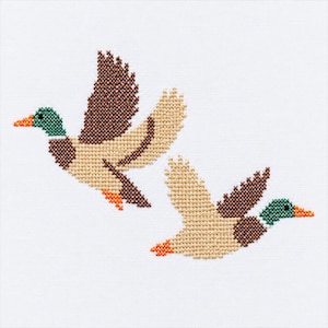 May include: A cross-stitch embroidery pattern on white fabric featuring two mallard ducks in flight. The ducks are rendered with dark green heads, orange beaks, brown wings, and light tan bodies, captured in a detailed pixelated stitch style.