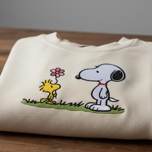 May include: Cream-colored sweatshirt featuring an embroidered design of Snoopy and Woodstock. Snoopy is depicted in black, white, and red, while Woodstock is yellow. The design includes a pink flower and green grass.