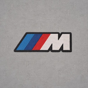 May include: A gray patch featuring the BMW M logo. The logo is a stylized "M" in white, with blue, dark blue, and red stripes on the left side, all outlined in black. The patch is on a gray textured background.