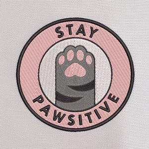 May include: Embroidered patch with a pink and black circular design. The text "STAY PAWSITIVE" surrounds a gray cat paw print. The patch is on a white background.