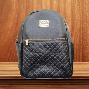 May include: Black leather backpack with a quilted design on the front pocket. The backpack has a gold zipper and a gold metal logo that says "C.F. Collection Design".