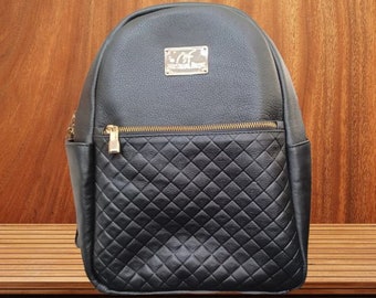 May include: Black leather backpack with a quilted design on the front pocket. The backpack has a gold zipper and a gold metal logo that says "C.F. Collection Design".
