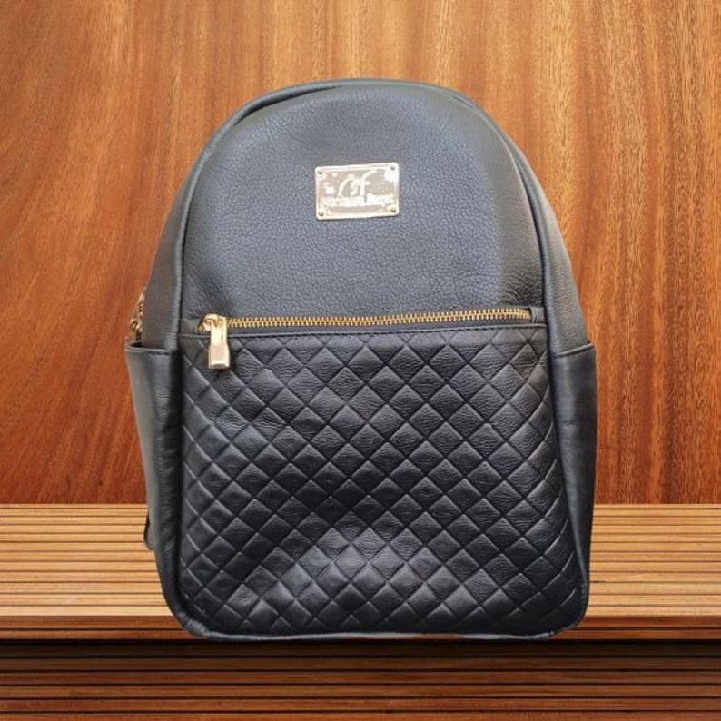 May include: Black leather backpack with a quilted design on the front pocket. The backpack has a gold zipper and a gold metal logo that says "C.F. Collection Design".