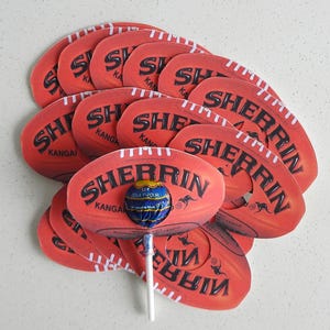 May include: A stack of red and white Australian Rules Football shaped lollipops with the word "SHERRIN" printed on them. The lollipops have a hole in the centre and a lollipop stick is inserted through the hole. The lollipops are decorated with white stitching and a kangaroo logo. The lollipop is a cola flavour.