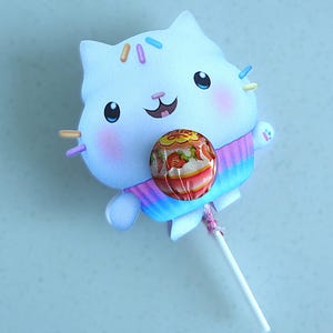 Puede incluir: Un soporte para piruletas con forma de gato blanco con una camisa a rayas de arcoíris de colores. El gato tiene mejillas rosadas, ojos azules y una cara sonriente. La piruleta está envuelta en un envoltorio transparente con un diseño de fresa.