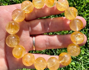 May include: A bracelet made of large, round, translucent citrine beads. The beads are a warm, golden-yellow color and appear to be polished. The bracelet is held in a hand against a background of green grass.