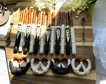 Puede incluir: Una colección de palitos y pretzels cubiertos de chocolate dispuestos sobre una tabla de madera. Algunos palitos están decorados como esmóquines con glaseado negro y pajaritas, mientras que otros tienen glaseado blanco y perlas de azúcar.