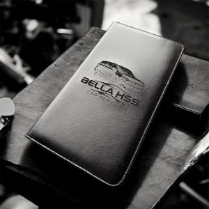 May include: Black leatherette car service folder with the text "BELLA HSS CAR SERVICE" and a car graphic. The folder is rectangular and has a stitched border. The image is in black and white.